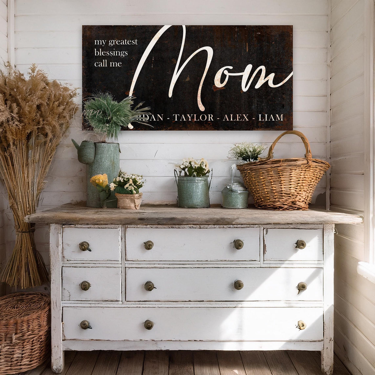 May include: A black and white sign with the word "Mom" in a cursive font. The sign also includes the text "My greatest blessings call me" and the names "Jordan, Taylor, Alex, Liam". The sign is hanging on a white wall above a white dresser with six drawers.